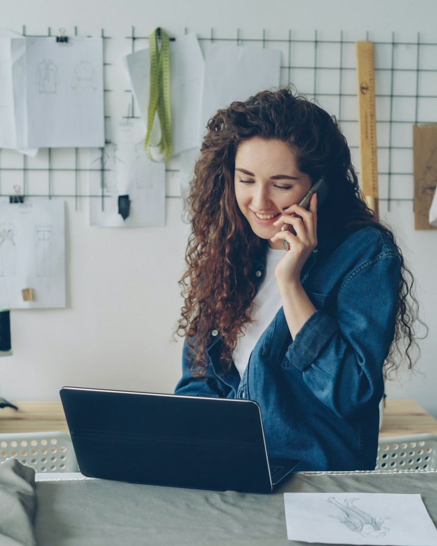 A fashion start-up founder on the phone at her laptop with a sewing machine and garments behind her