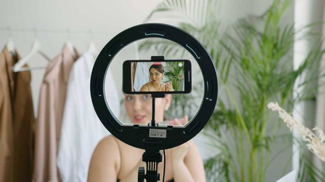 A fashion content creator filming herself on a phone in a ring light, with a rack of gifted clothing behind her