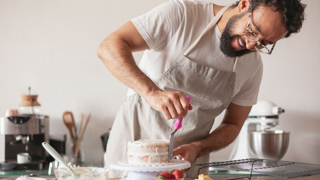 A home baker in a linen apron smoothing icing onto a layer cake on a turntable at his kitchen bench, with strawberries and a stand mixer nearby