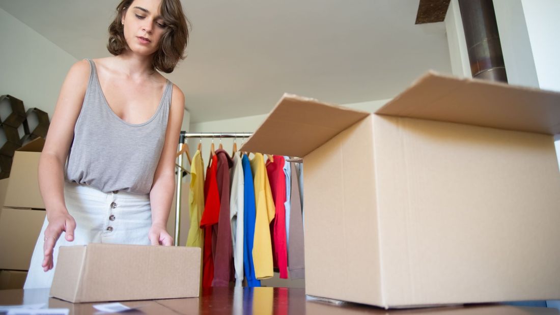A young clothing-store founder packing a box for a restock order, with a rail of brightly coloured garments behind her
